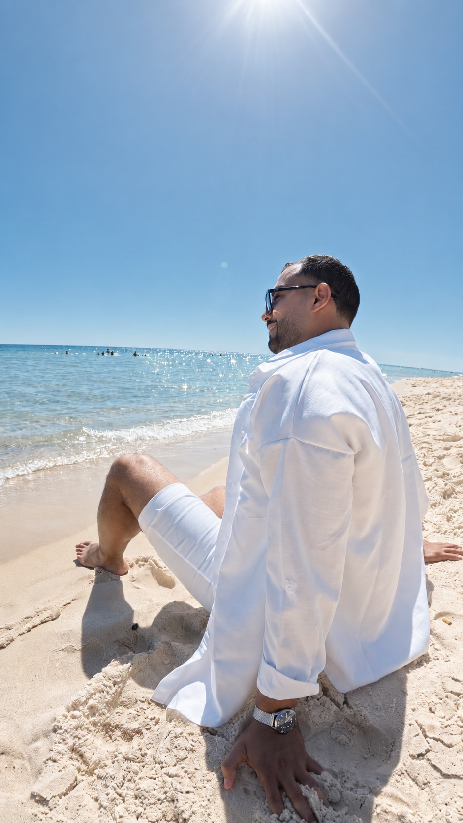 Man in white shirt and shorts sitting on a sandy beach with ocean and clear blue sky in the background.