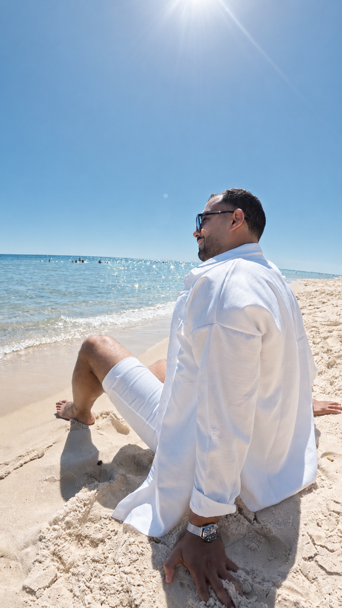 Man in white shirt and shorts sitting on a sandy beach with ocean and clear blue sky in the background.